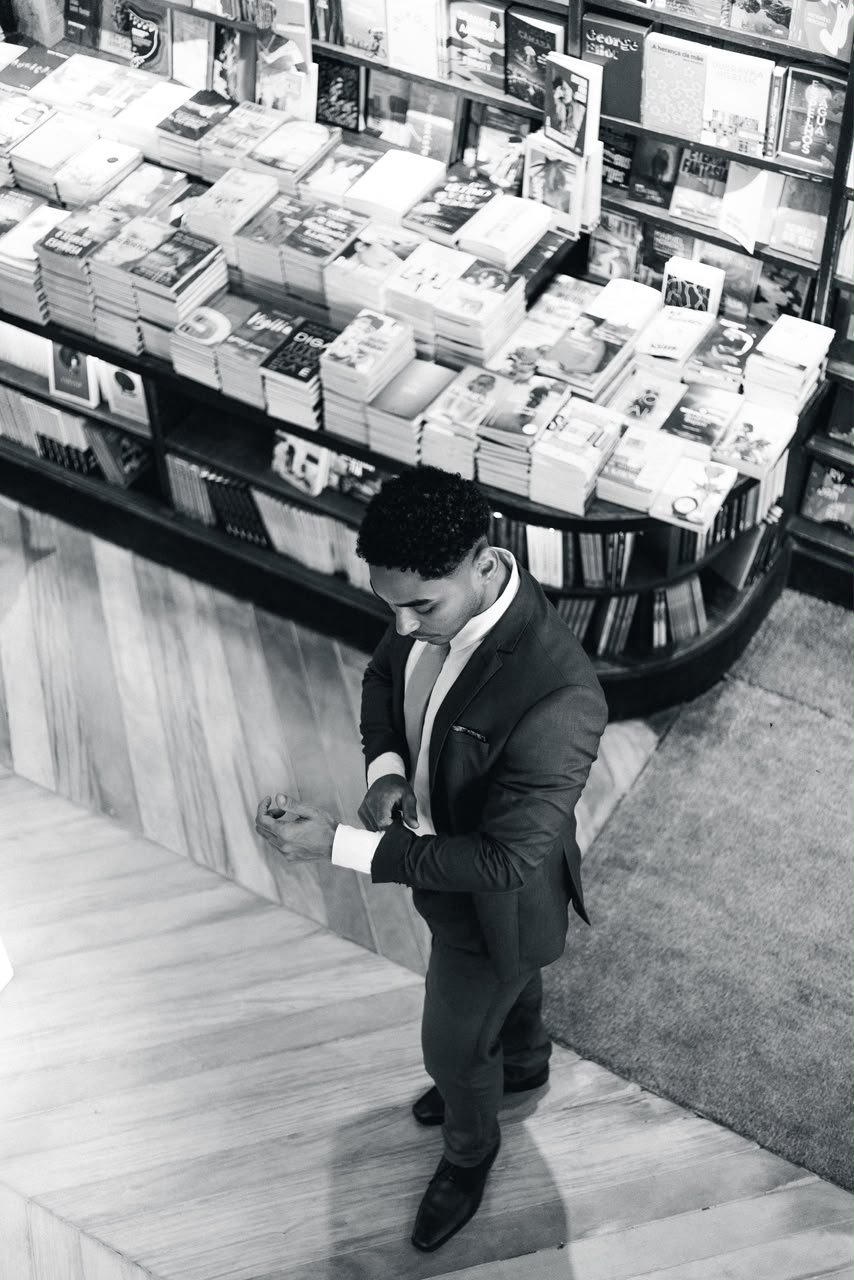 Foto em preto e branco de Douglas Aleixo de corpo inteiro, ajustando o punho da camisa em uma livraria